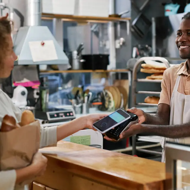 A woman holding a bag of bread at a bakery pays at the POS terminal using her smartphone.